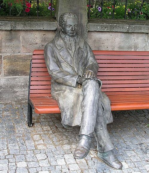Das Bild zeigt das Goethe-Denkmal am Ilmenauer Markt, eine Bronzefigur auf einer Holzbank sitzend.
