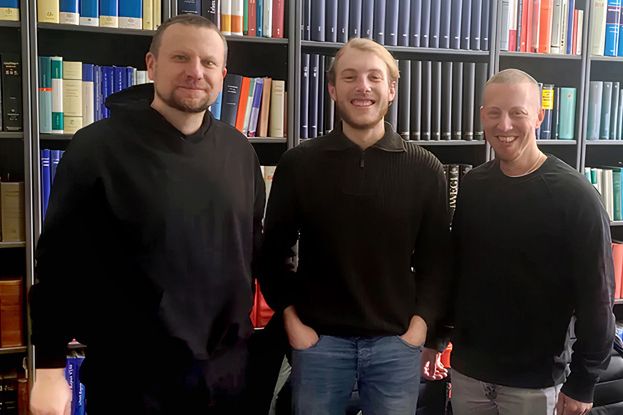Ein Foto vom dreiköpfigen Team des Gaming-Studios Inclusive Gaming, vor einer Bücherwand stehend.