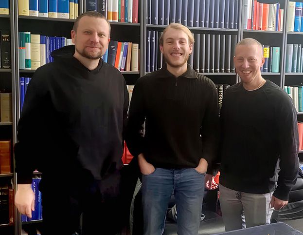 Ein Foto vom dreiköpfigen Team des Gaming-Studios Inclusive Gaming, vor einer Bücherwand stehend.